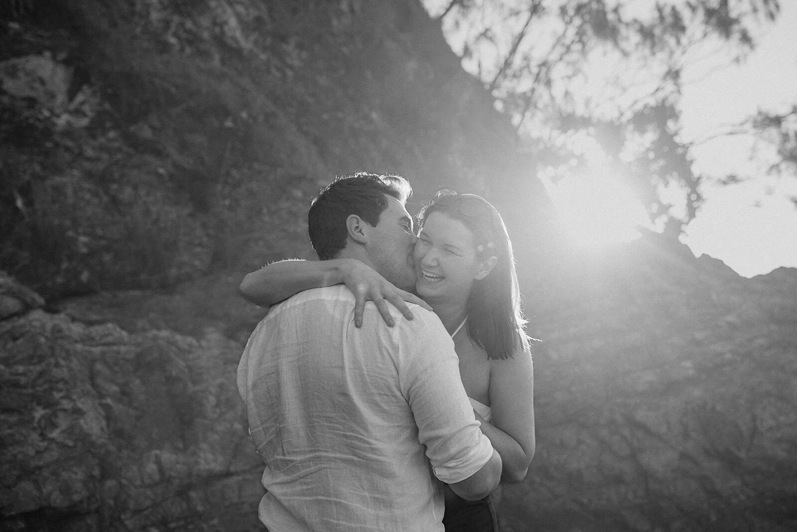 Black and white of the couple laughing and embracing in the backlight, John and Emma's sunset marriage proposal at Wategos Beach, Byron Bay