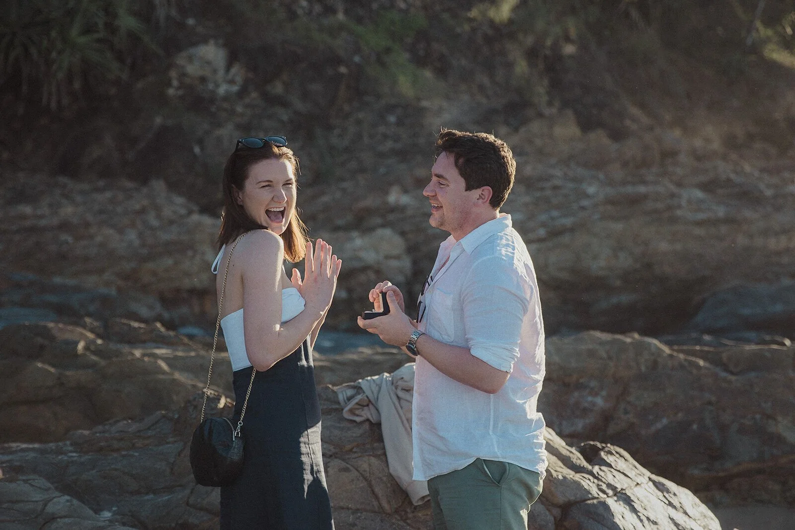 Woman laughing with her hand raised as her partner holds the ring box, John and Emma's sunset marriage proposal at Wategos Beach, Byron Bay