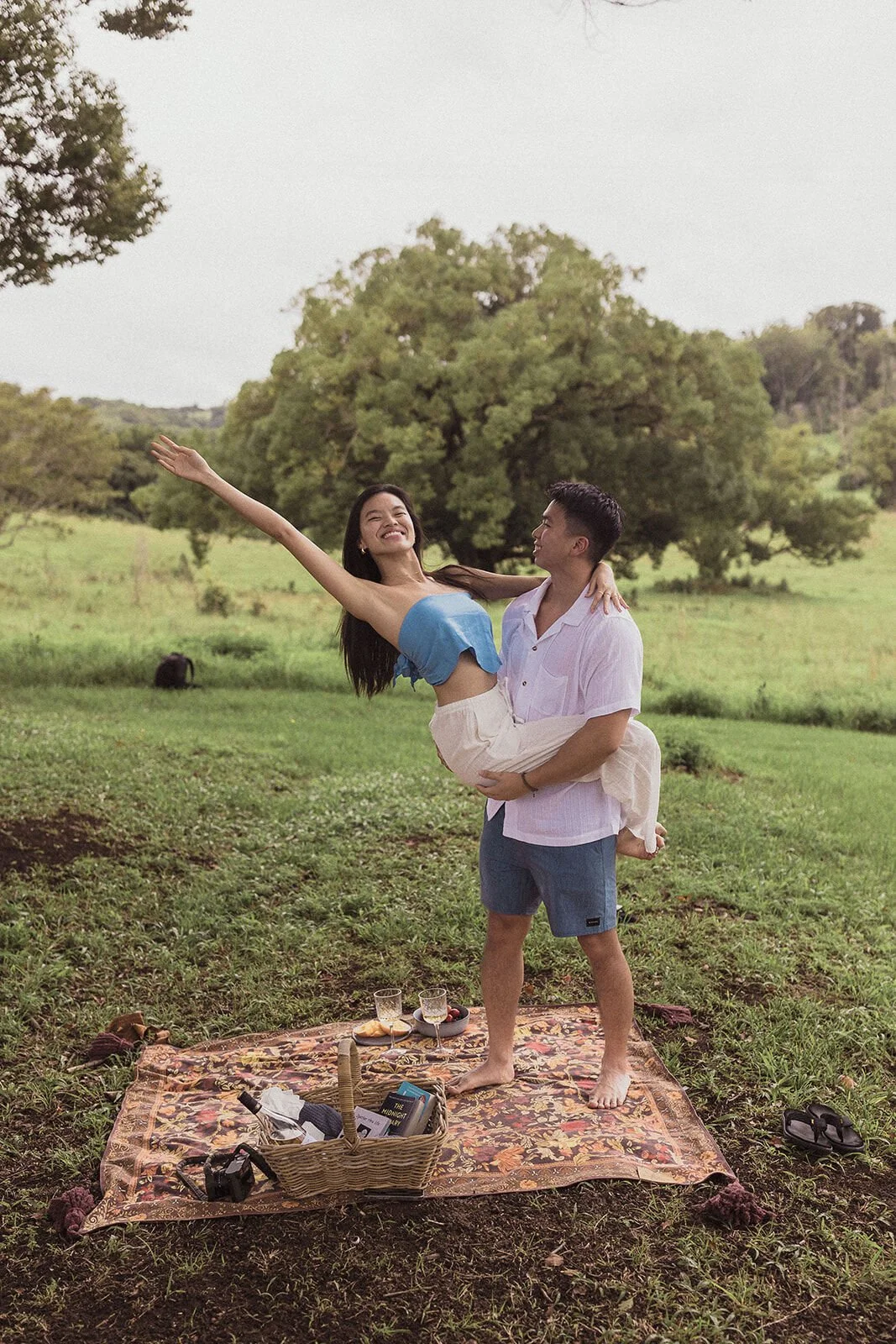 Man holding his fiancée in his arms with her arm outstretched beside the picnic rug, Darien and Jacqui's picnic marriage proposal at Heartwood Farm, Byron Bay hinterland