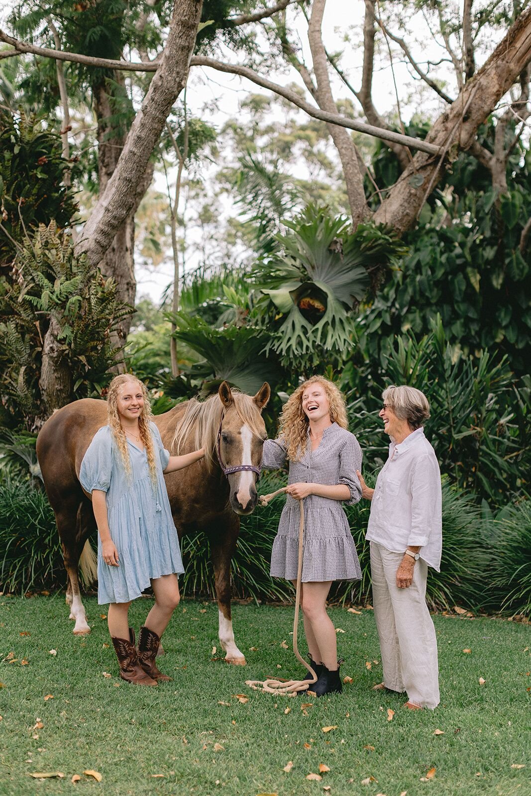 Two sisters in blue dresses laughing with their grandmother and a horse under tall trees