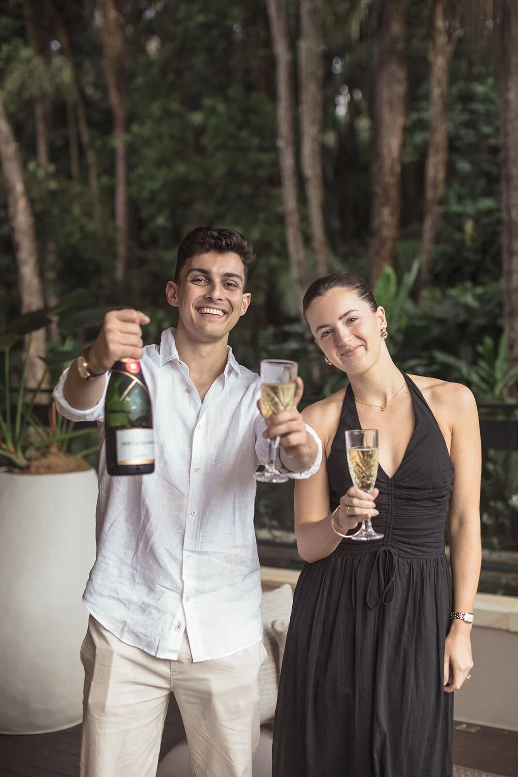 Couple raising the champagne bottle and glasses to the camera, Ben and Chloe's marriage proposal in Byron Bay