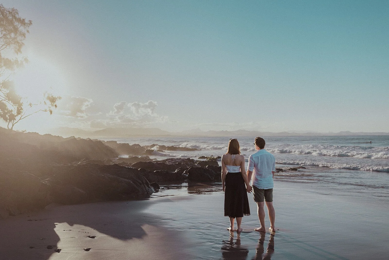 Couple from behind holding hands and looking out to sea, John and Emma's sunset marriage proposal at Wategos Beach, Byron Bay