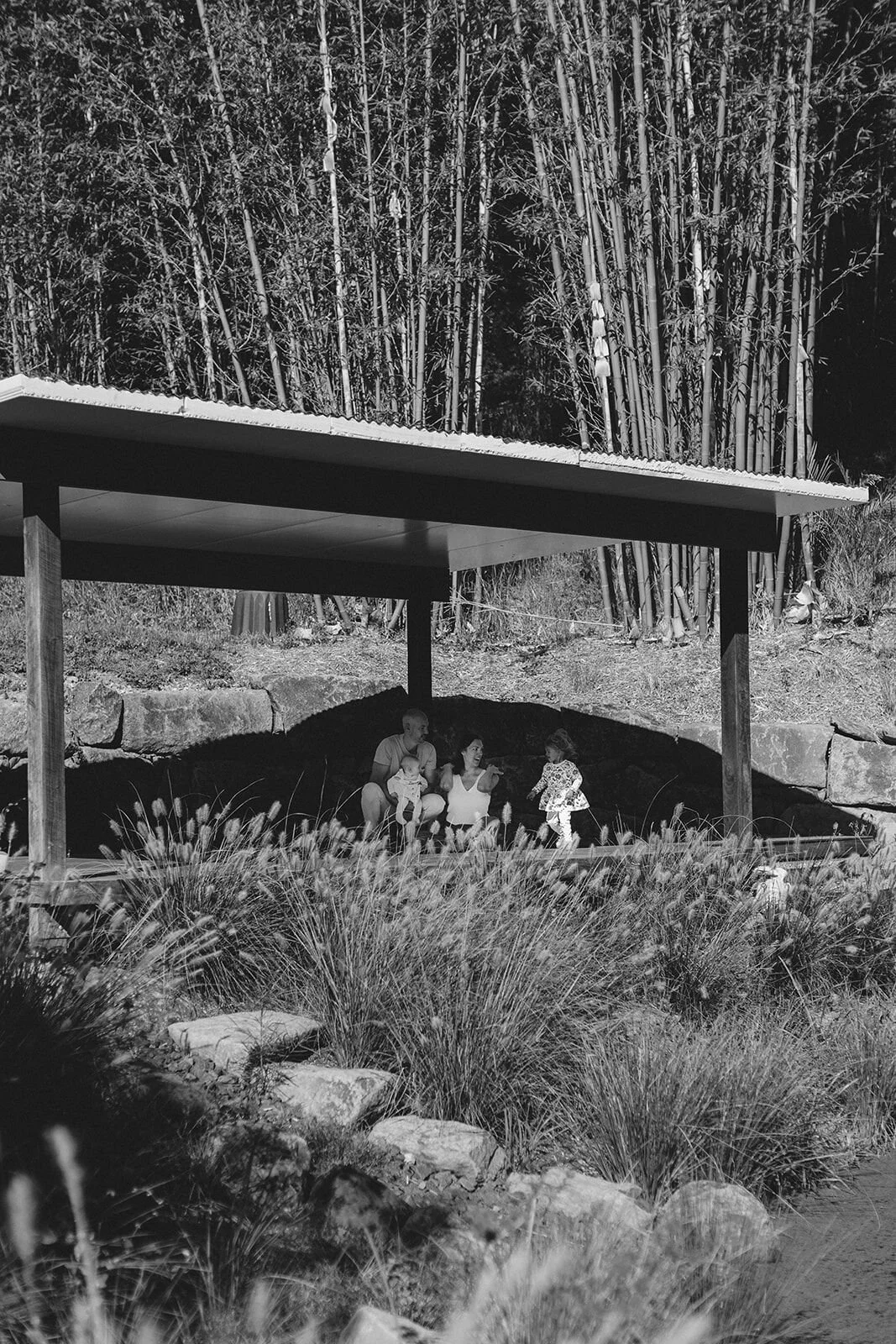 Black and white wide shot of a family sitting under a timber pergola with a bamboo grove behind