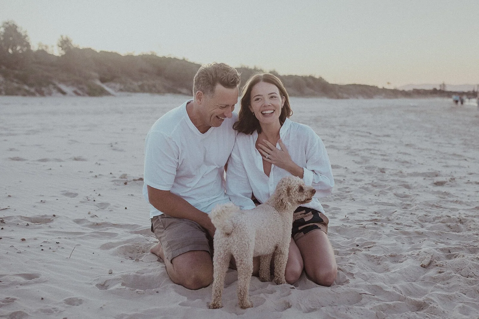 Woman with her hand over her mouth smiling beside her fiancé and their dog, Jamie and Jessica's beach marriage proposal at Belongil Beach, Byron Bay