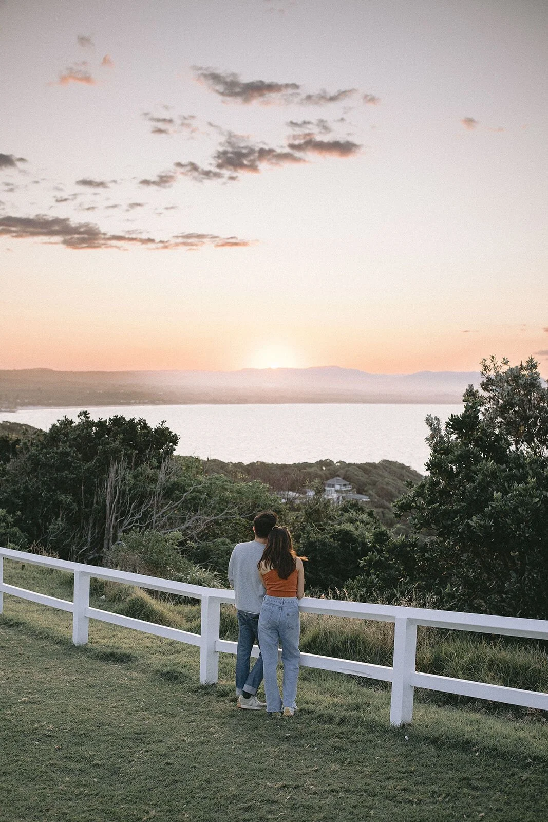 Couple at the white fence watching the sun go down over the water, Douglas and Ying's sunset marriage proposal at Cape Byron Lighthouse, Byron Bay