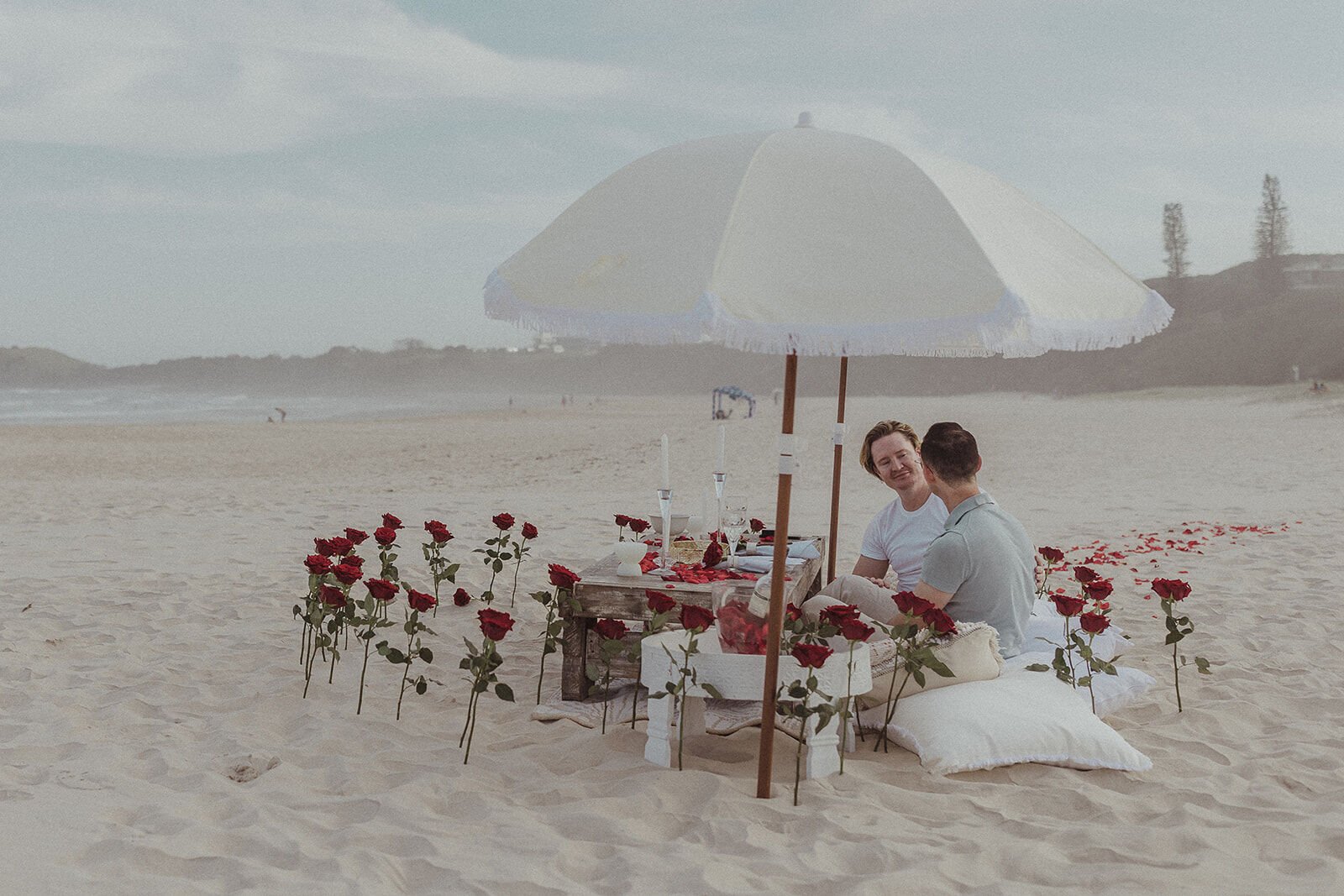 Couple chatting at the rose-lined beach picnic under a white umbrella, Byron and Luke's marriage proposal at Cabarita Beach, Northern Rivers