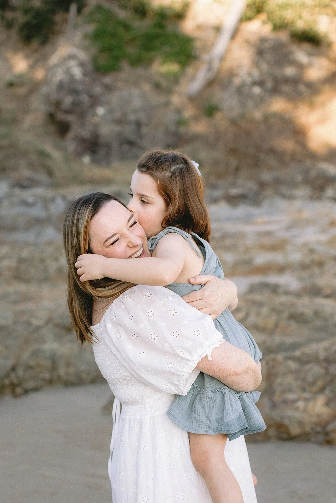Mum hugging her daughter close on the beach, the little girl kissing her cheek