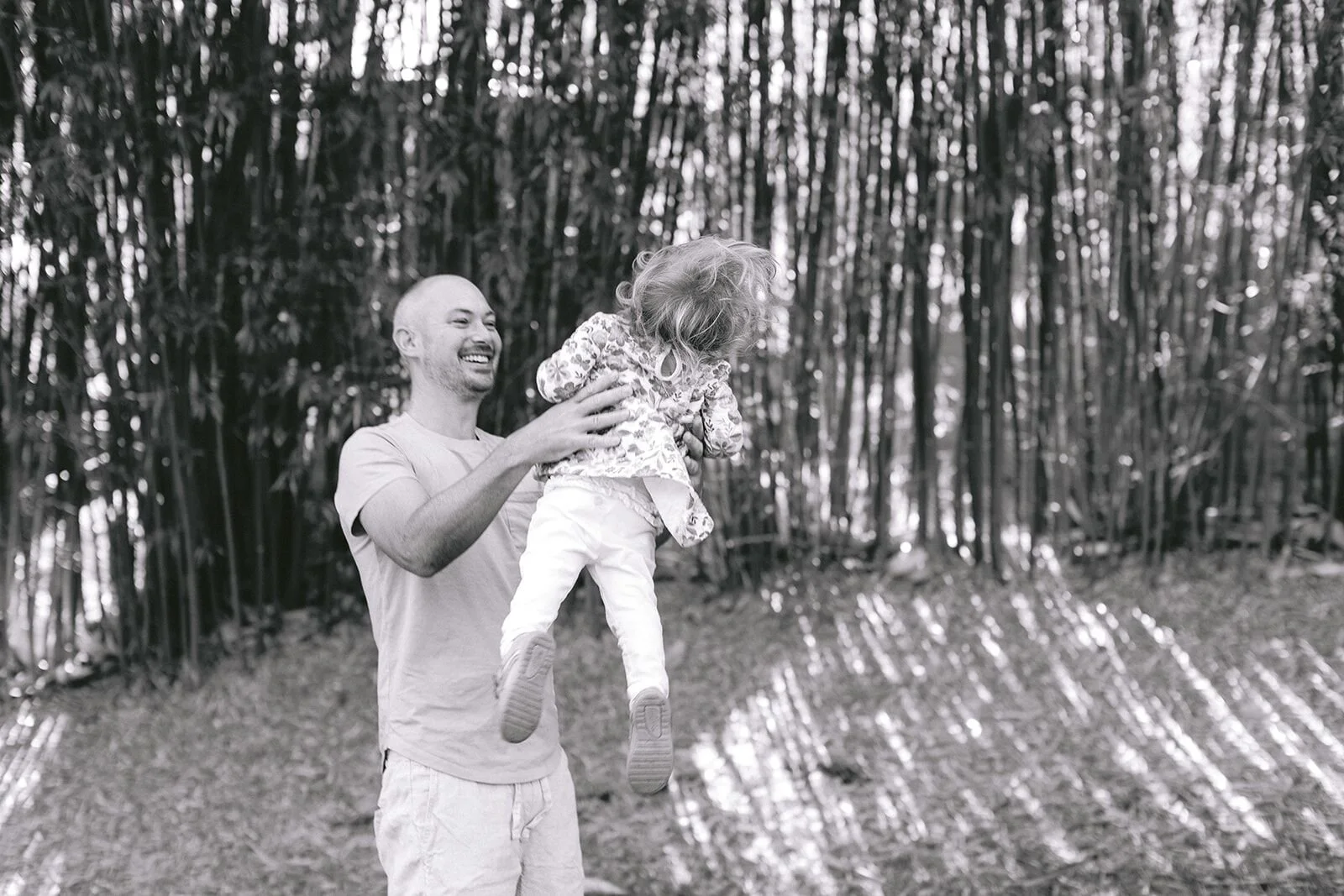 Black and white photo of dad lifting his toddler daughter, both laughing, bamboo grove behind