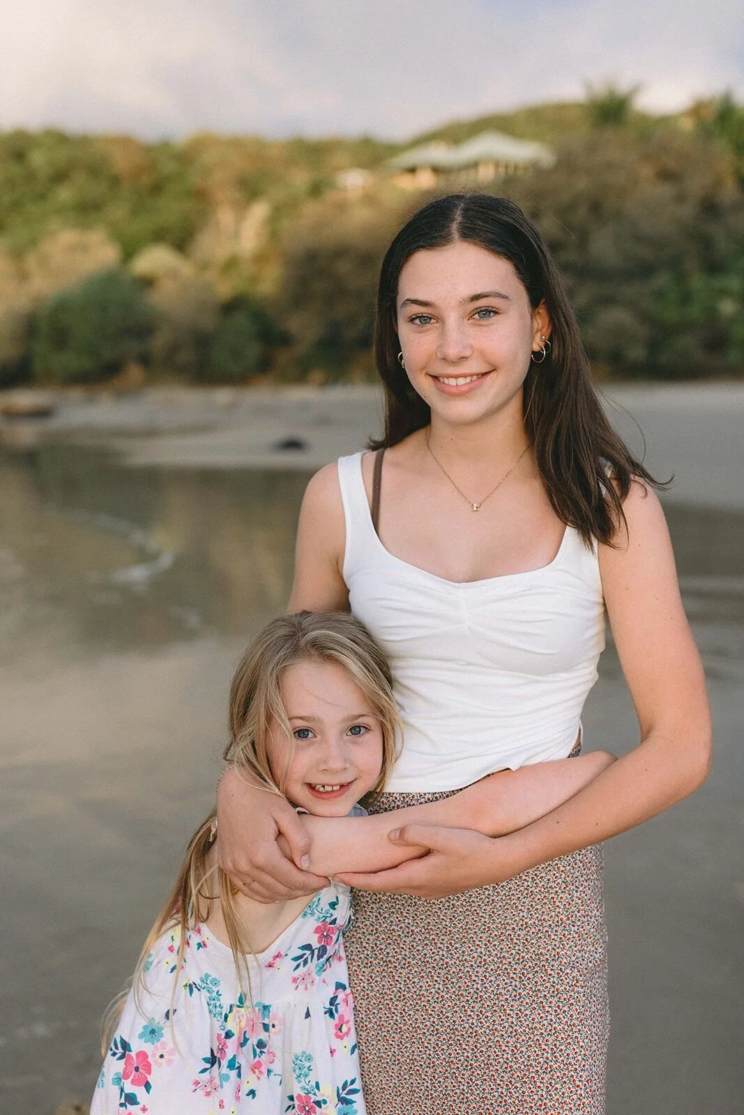 Teenager hugging her little sister from behind on the sand, both smiling