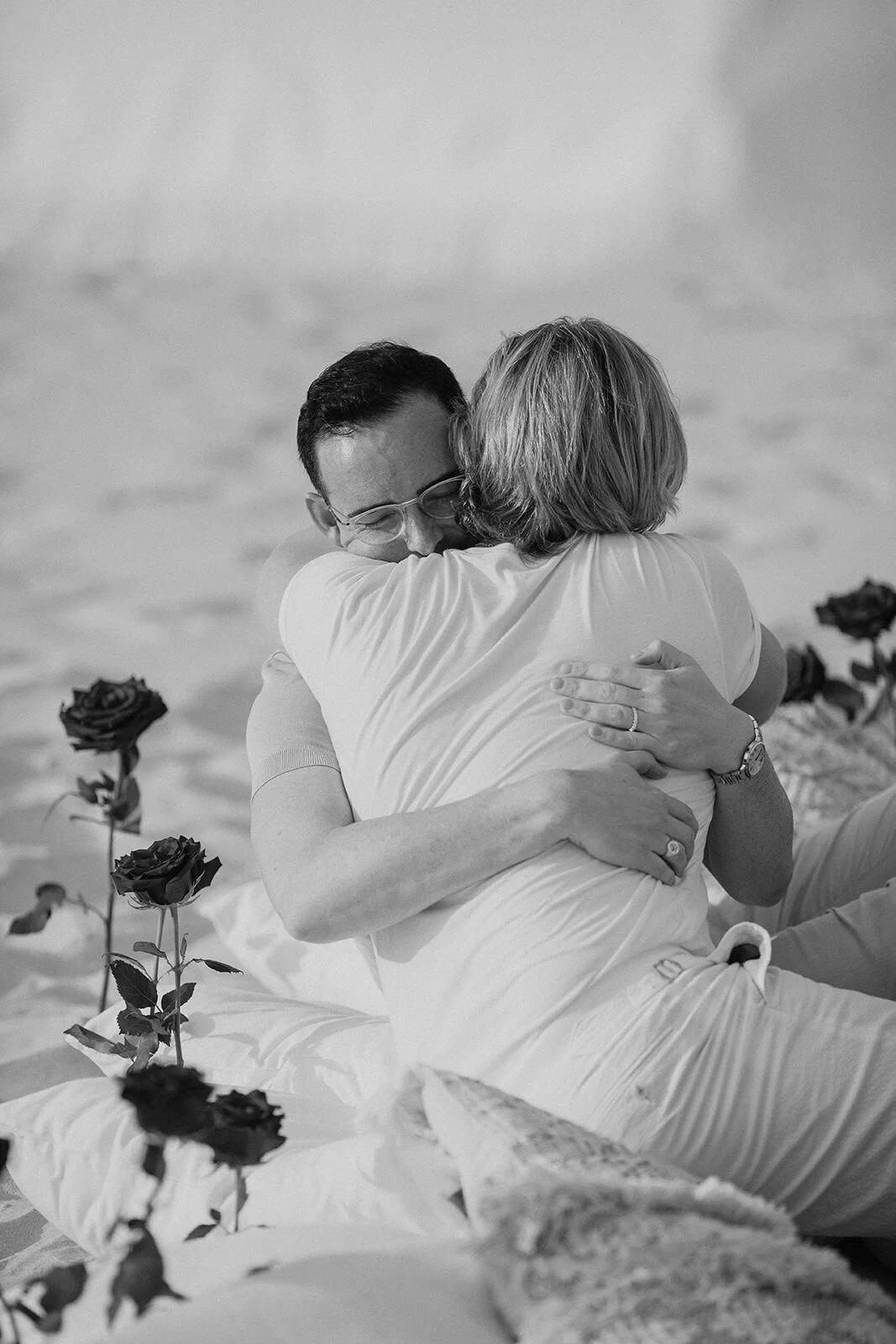 Black and white of the couple embracing among the red roses on the sand, Byron and Luke's marriage proposal at Cabarita Beach, Northern Rivers