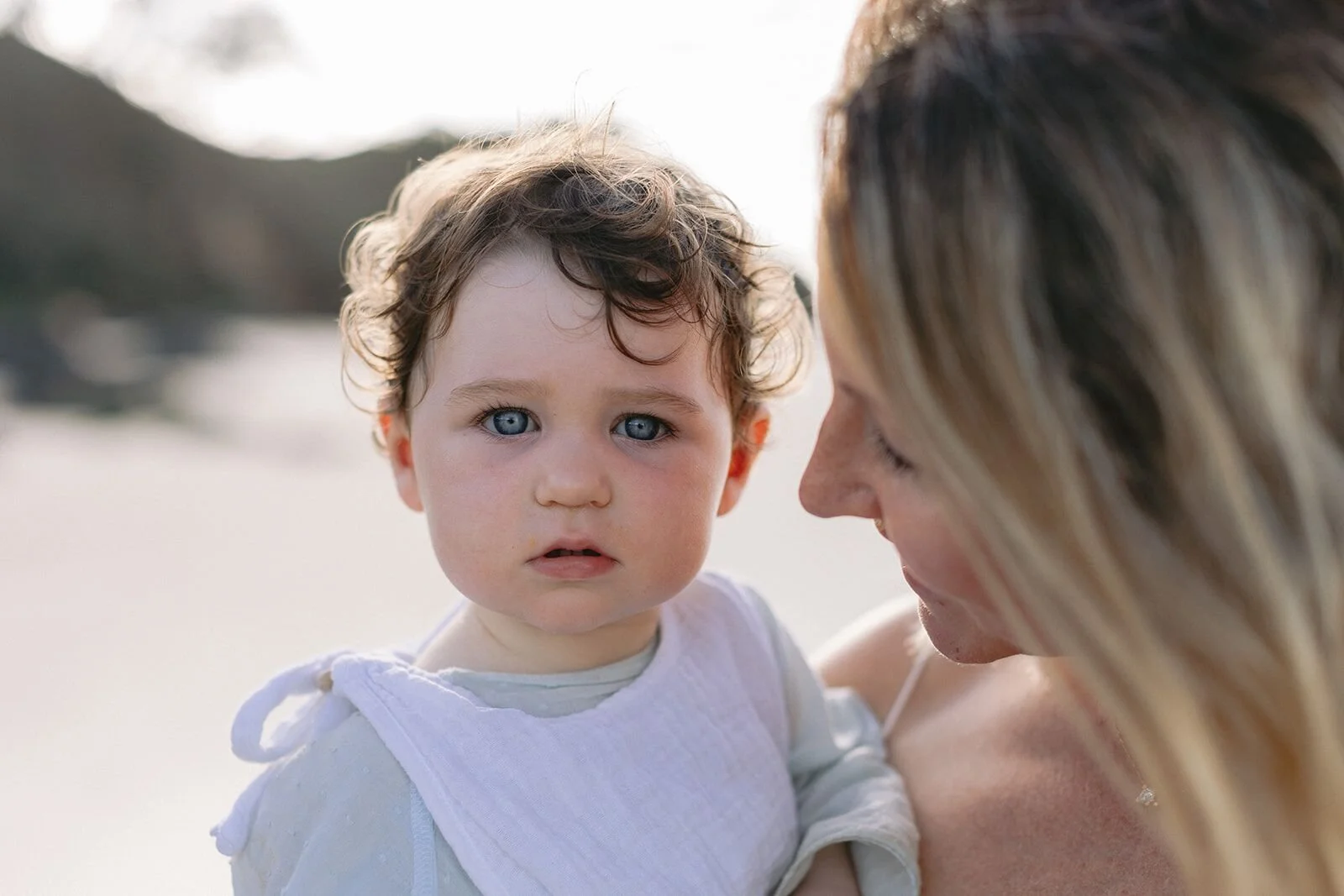 Close-up of a blue-eyed toddler in his mum's arms at Wategos Beach in soft evening light