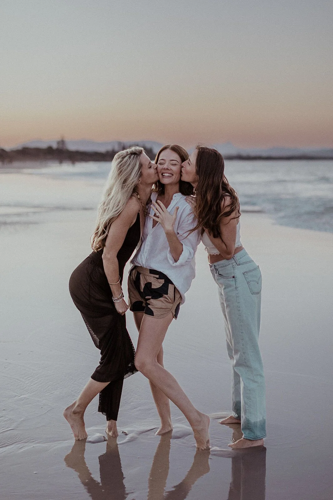 Two friends kissing the woman on the cheeks at the water's edge, Jamie and Jessica's beach marriage proposal at Belongil Beach, Byron Bay