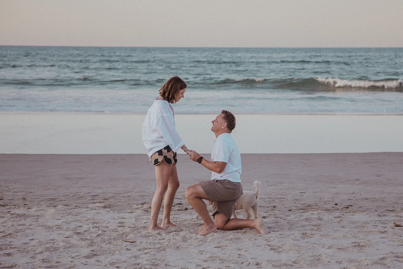 Man holding out the ring on one knee as the woman looks down in surprise, Jamie and Jessica's beach marriage proposal at Belongil Beach, Byron Bay