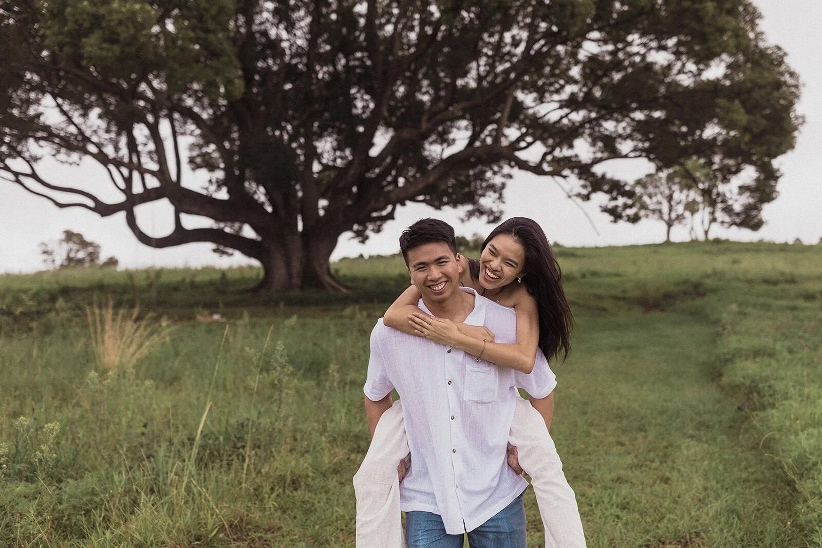 Man giving his fiancée a piggyback, both laughing in the field, Darien and Jacqui's picnic marriage proposal at Heartwood Farm, Byron Bay hinterland
