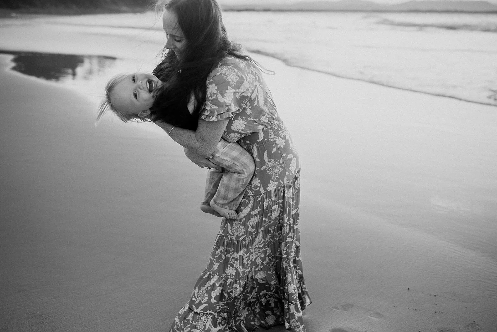 Black and white photo of a toddler giggling as mum swings him up on the sand