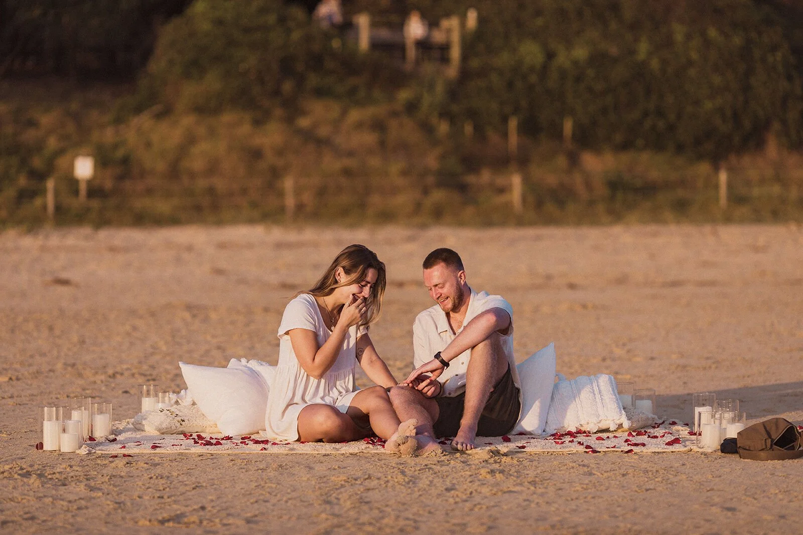 Couple laughing together and looking at the ring on the blanket, Charlie and Hannah's sunrise marriage proposal at Broken Head Beach, Byron Bay