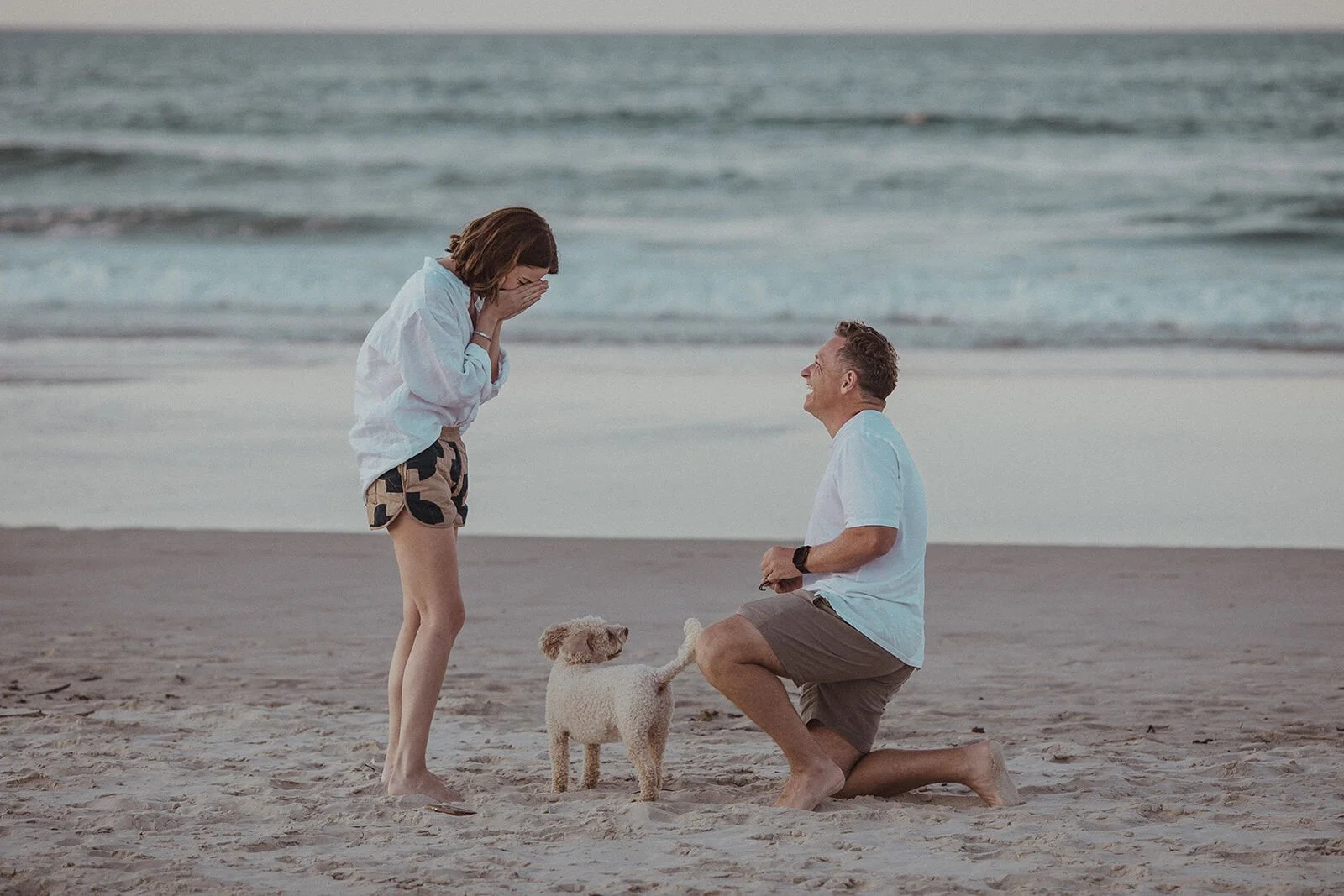Man kneeling on the sand to propose as the woman covers her mouth, their dog beside them, Jamie and Jessica's beach marriage proposal at Belongil Beach, Byron Bay