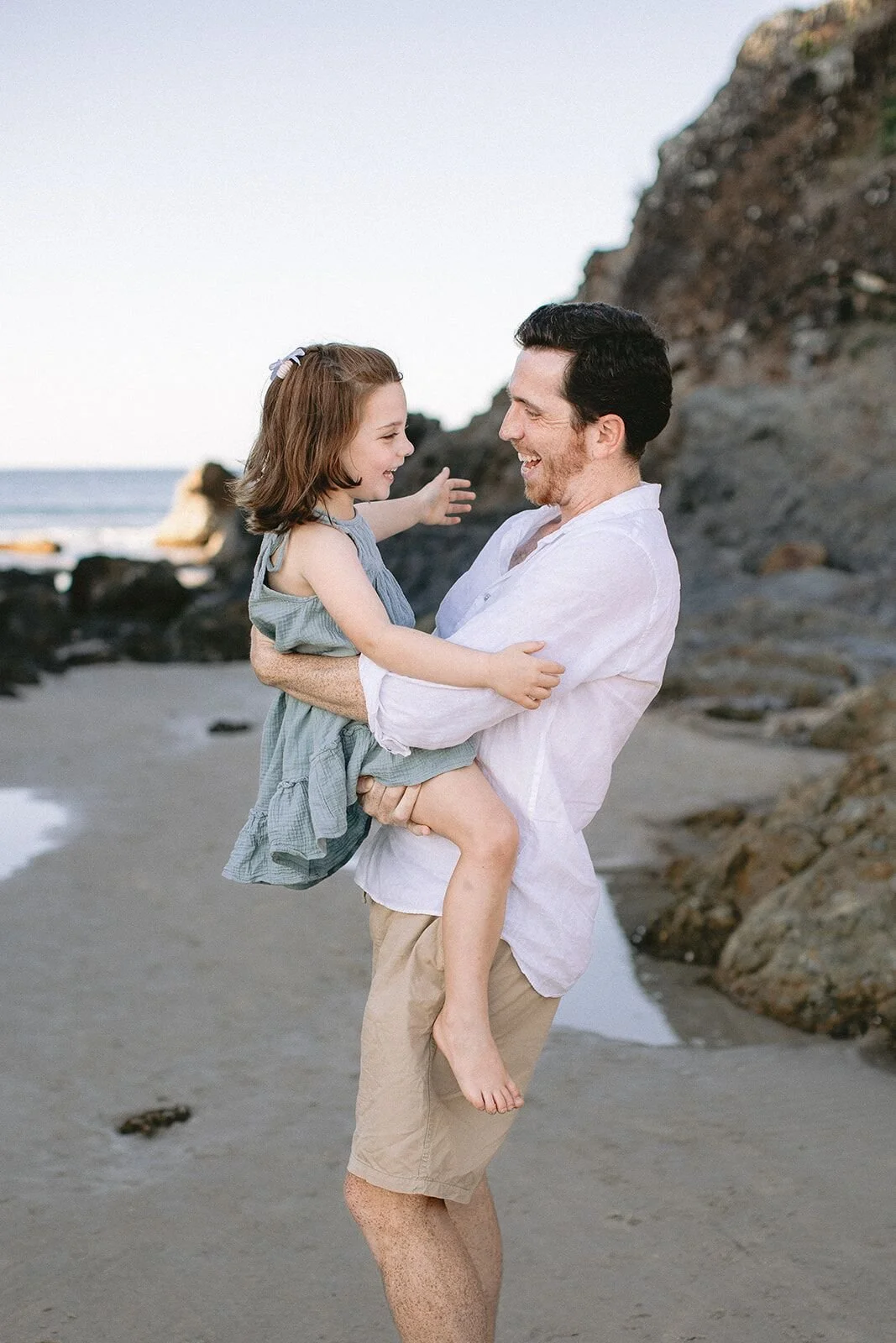 Dad in a white shirt carrying his daughter on the sand, both smiling