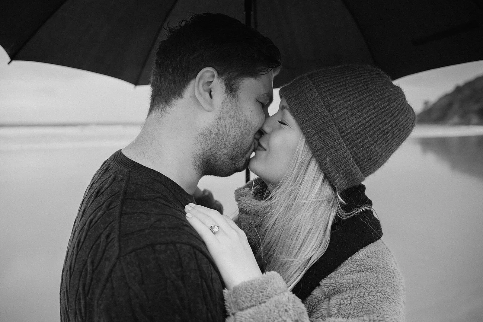 Black and white of the couple kissing under the umbrella, Raphael and Vany's rainy day marriage proposal at Broken Head Beach, Byron Bay