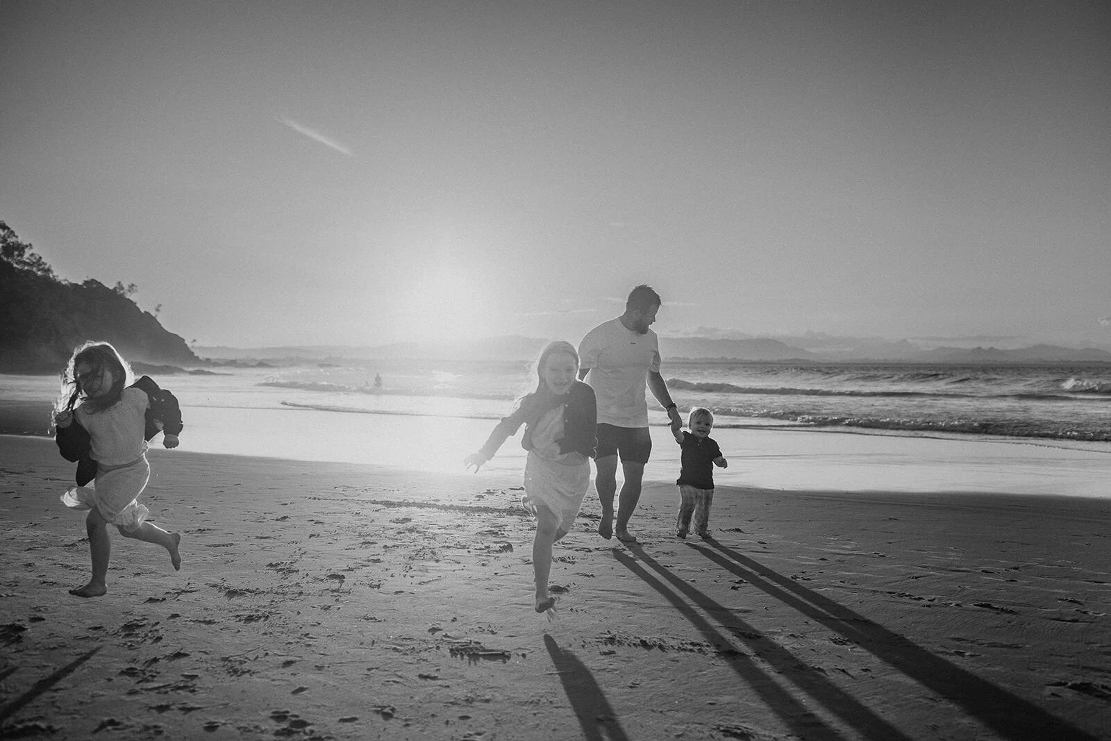 Black and white photo of dad walking with his toddler while his daughter runs ahead, long shadows on the sand