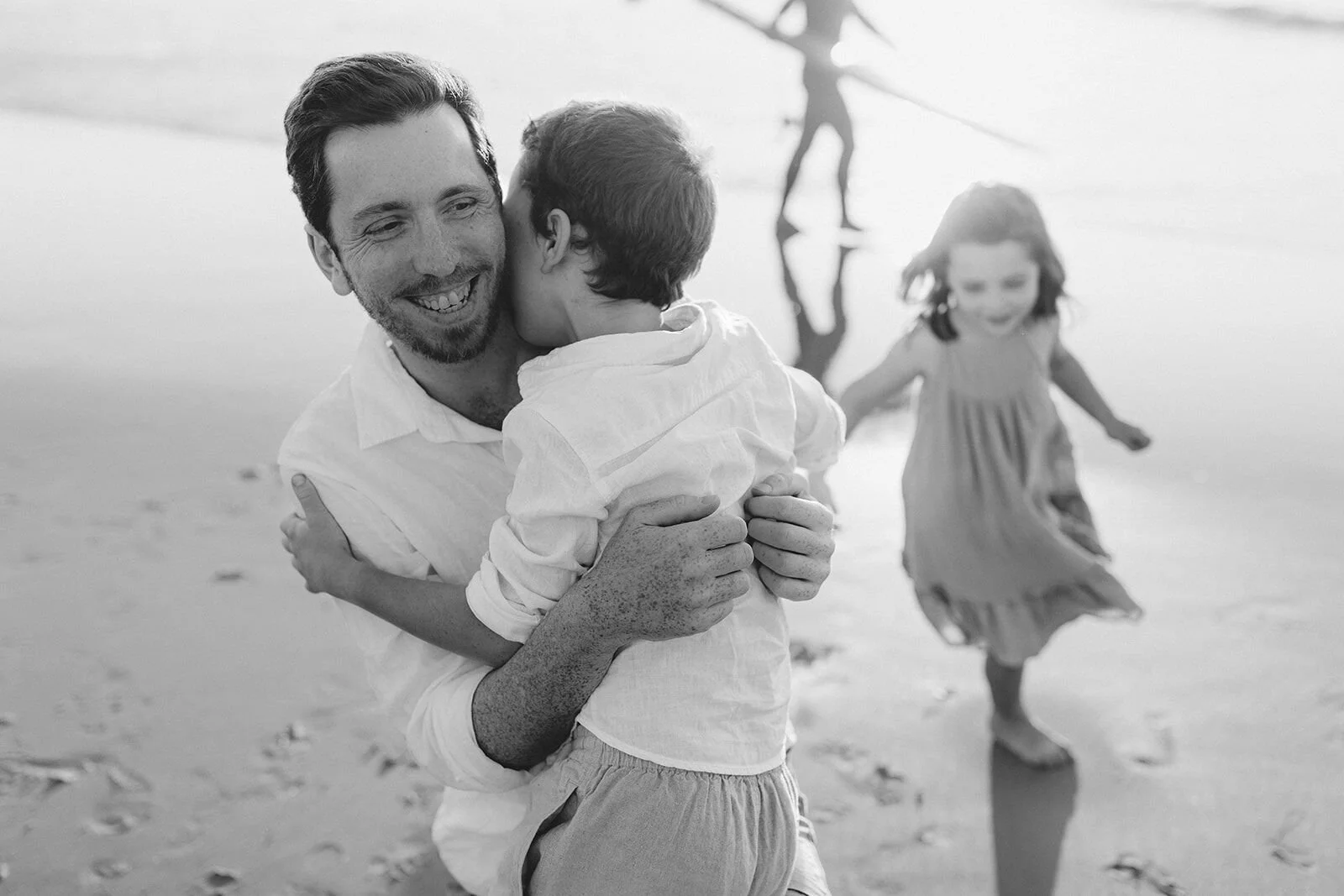 Black and white photo of dad hugging his son while his daughter runs past on the sand