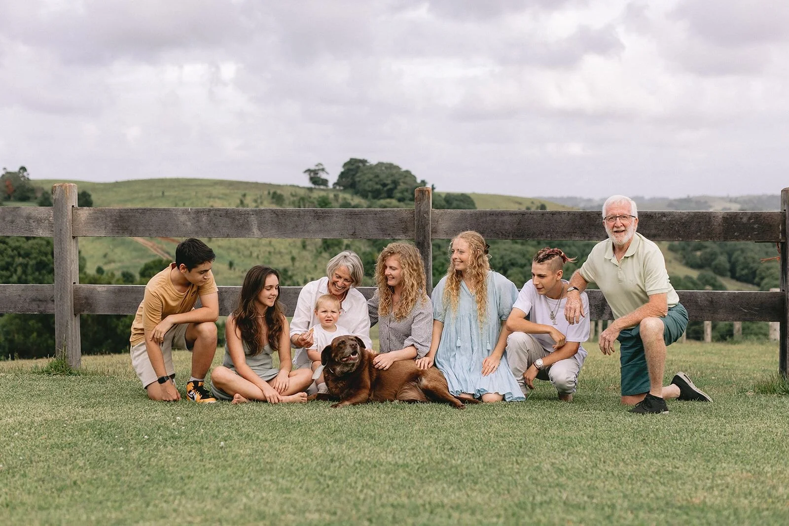 Family sitting on the grass in front of a farm fence with their chocolate Labrador