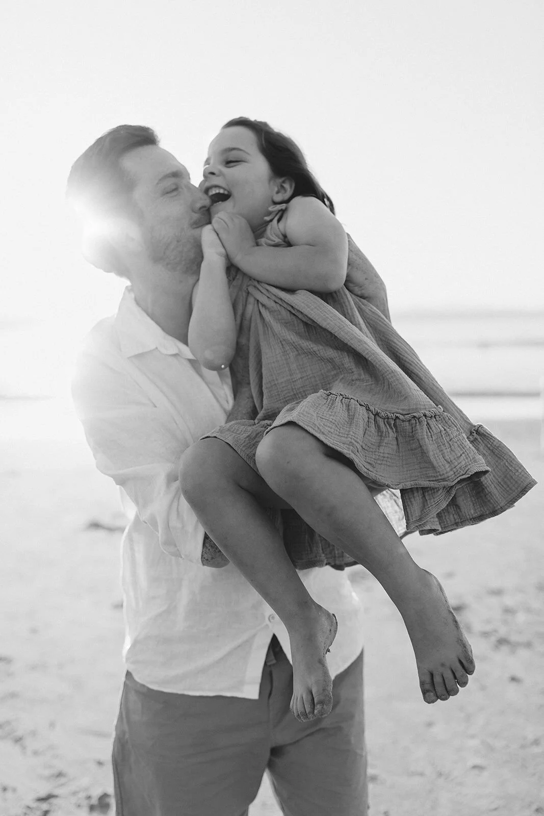 Black and white photo of dad kissing his giggling daughter as he carries her on the beach