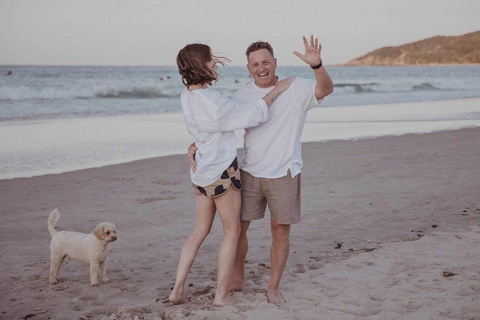 Couple laughing together as he waves, their dog beside them, Jamie and Jessica's beach marriage proposal at Belongil Beach, Byron Bay