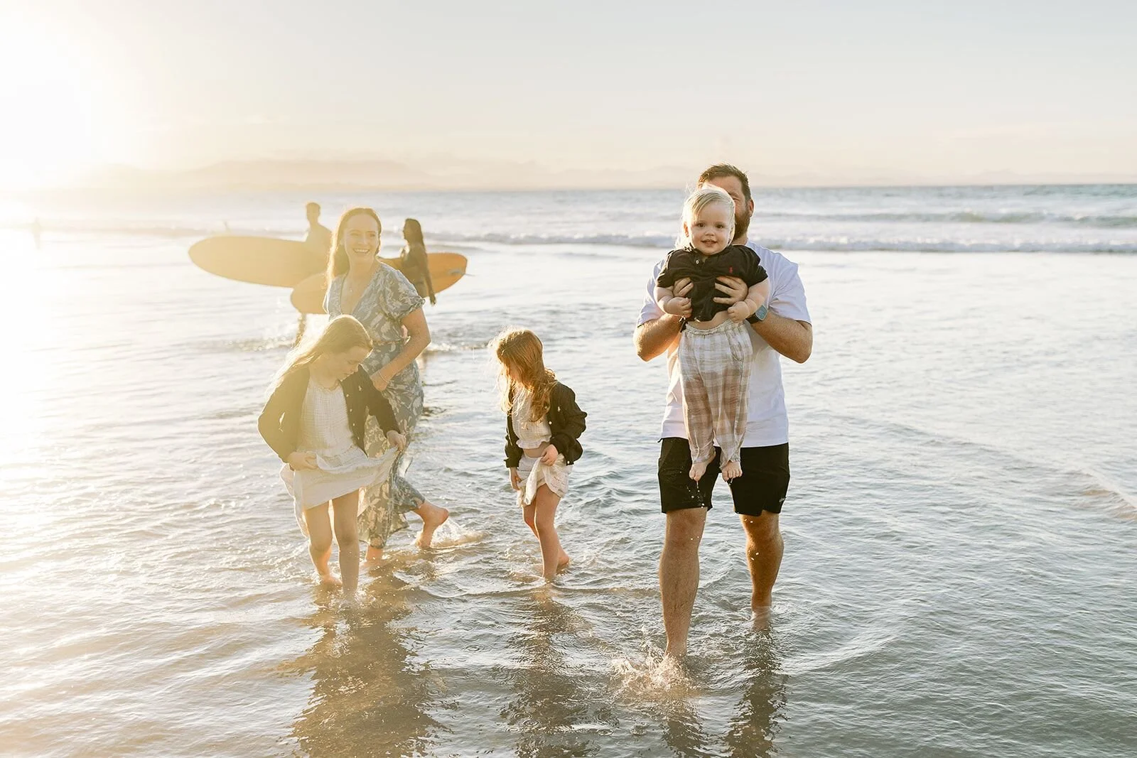 Family wading through the water at golden hour with a wooden surfboard in the background