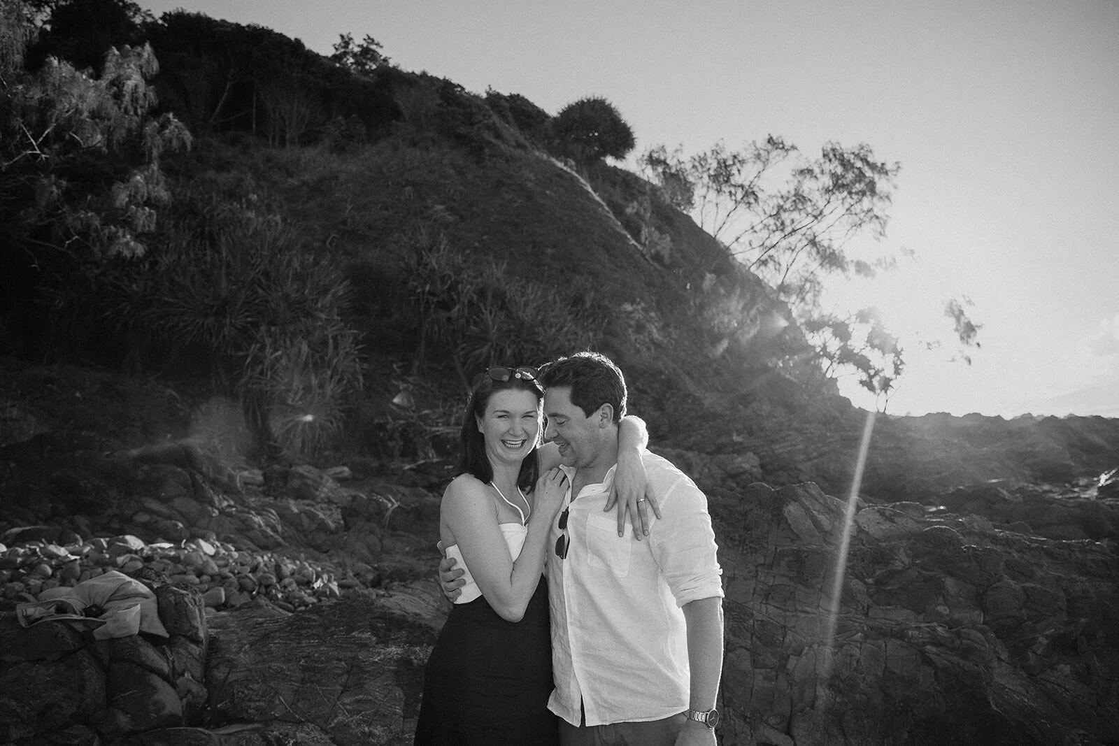 Black and white of the couple laughing with their arms around each other, John and Emma's sunset marriage proposal at Wategos Beach, Byron Bay