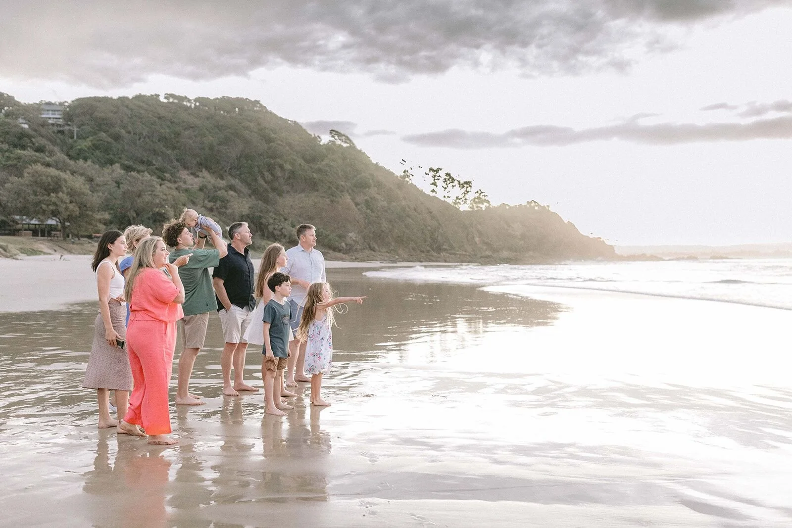 Extended family standing on the wet sand looking out to sea at sunset, the headland behind them