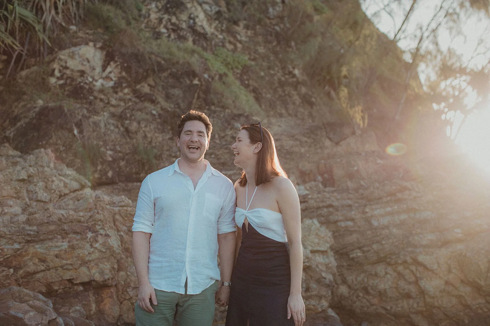 Couple laughing together on the rocks in the sunset light, John and Emma's sunset marriage proposal at Wategos Beach, Byron Bay