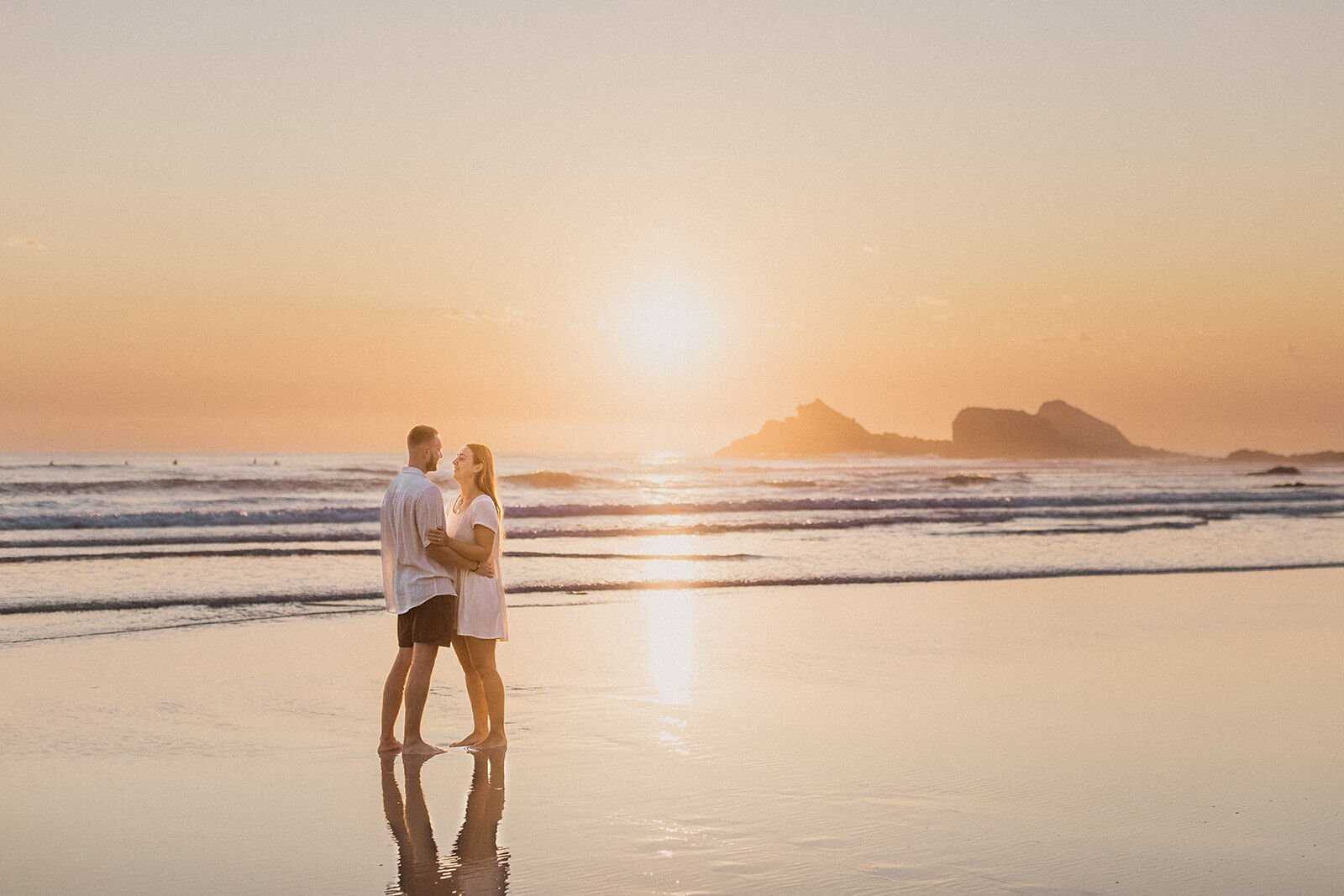 Couple embracing in the distance with the sea stacks and rising sun behind, Charlie and Hannah's sunrise marriage proposal at Broken Head Beach, Byron Bay