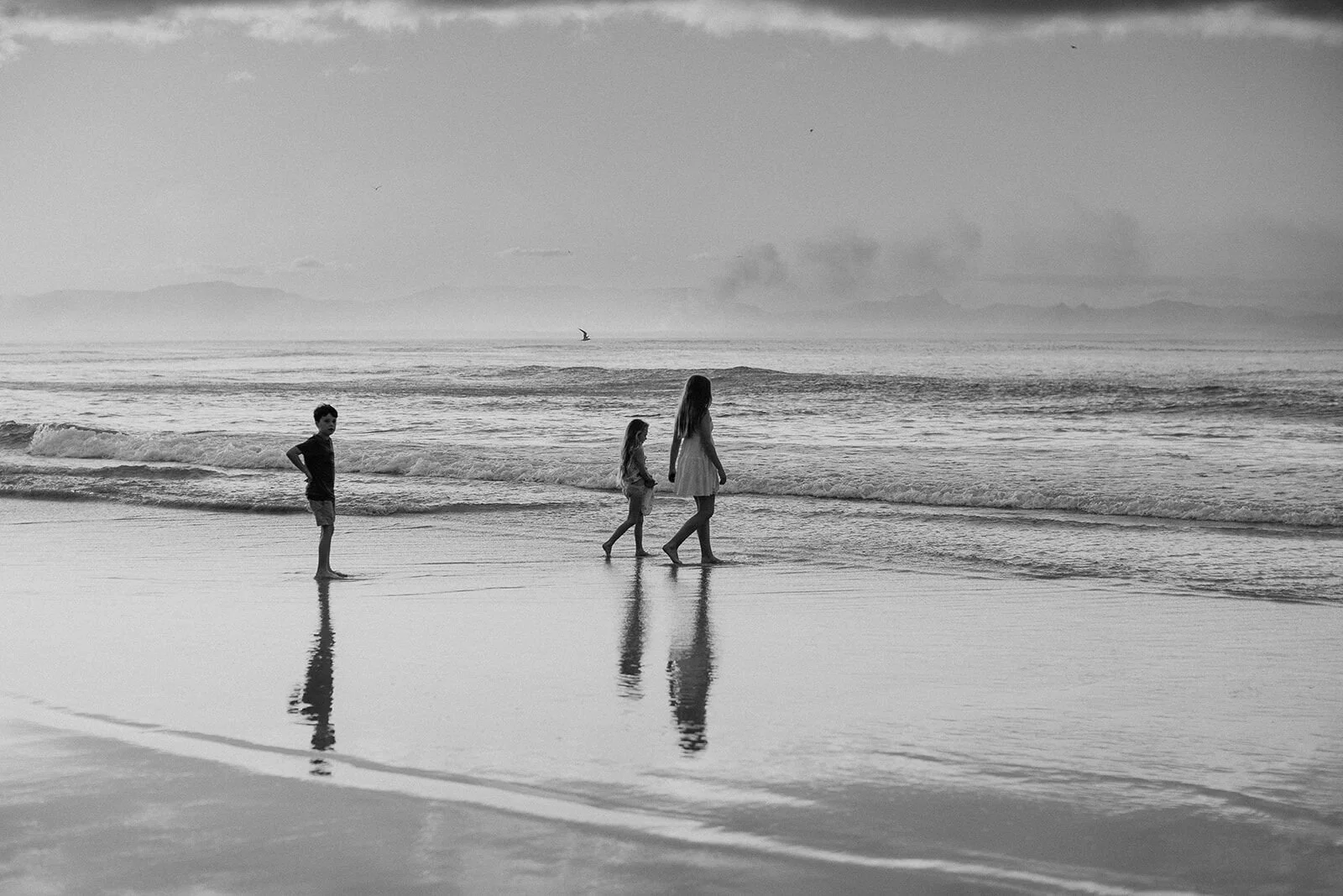 Black and white wide shot of children walking along the water's edge on an empty beach