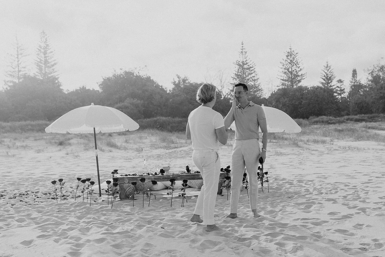 Black and white of the couple by the picnic as one wipes a happy tear, Byron and Luke's marriage proposal at Cabarita Beach, Northern Rivers