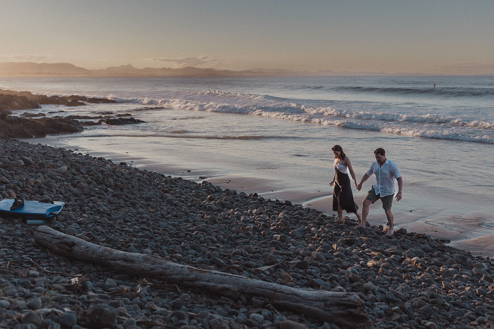 Couple walking hand in hand along the pebbly shoreline, John and Emma's sunset marriage proposal at Wategos Beach, Byron Bay