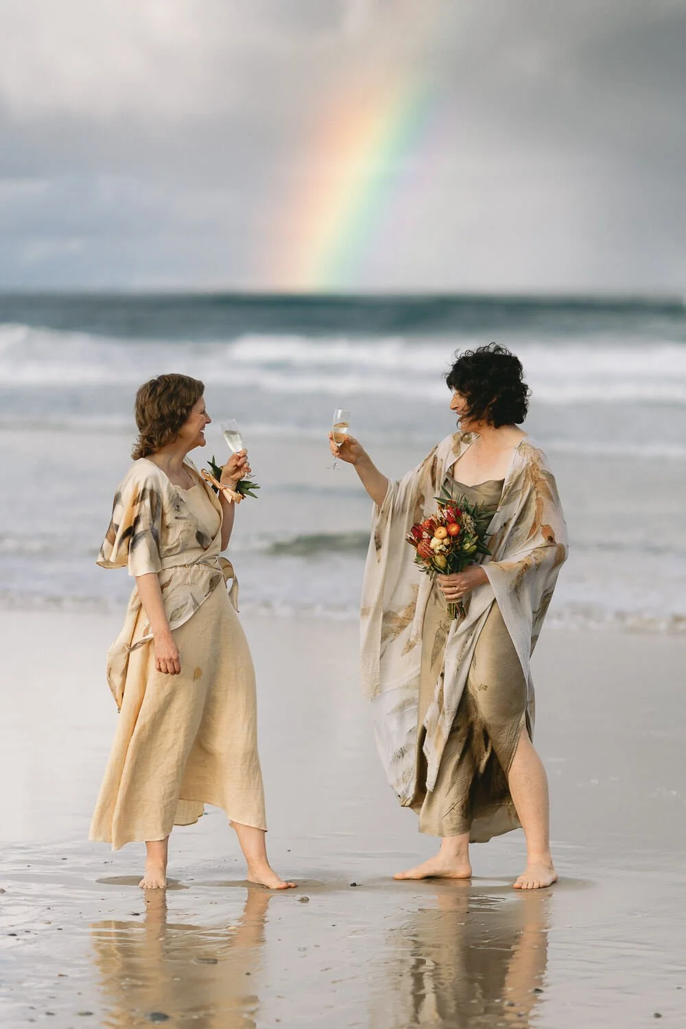 Two brides toasting with champagne under a rainbow on Seven Mile Beach, Lennox Head
