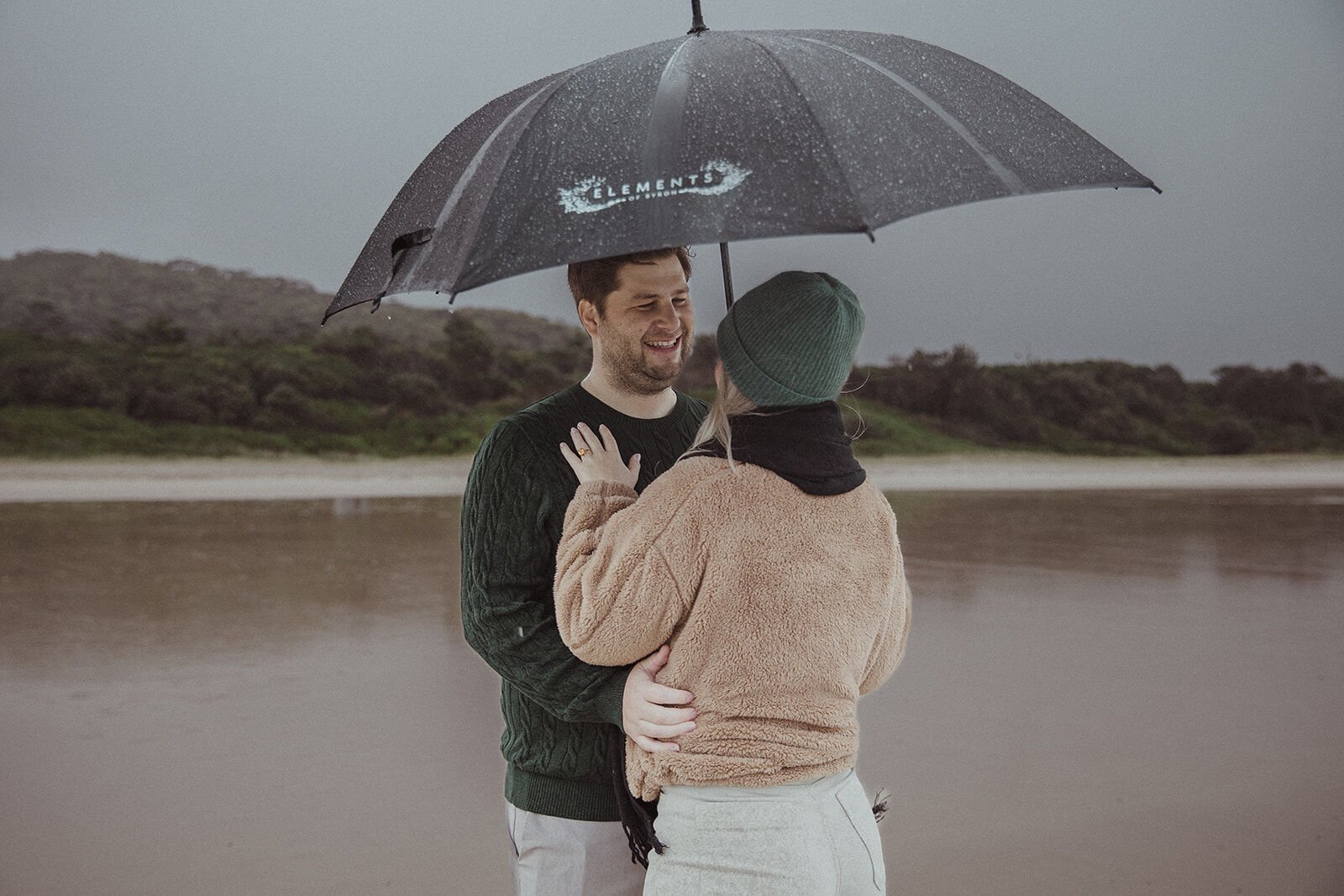 Man smiling as his fiancée embraces him, her ringed hand raised, beside the wet sand, Raphael and Vany's rainy day marriage proposal at Broken Head Beach, Byron Bay