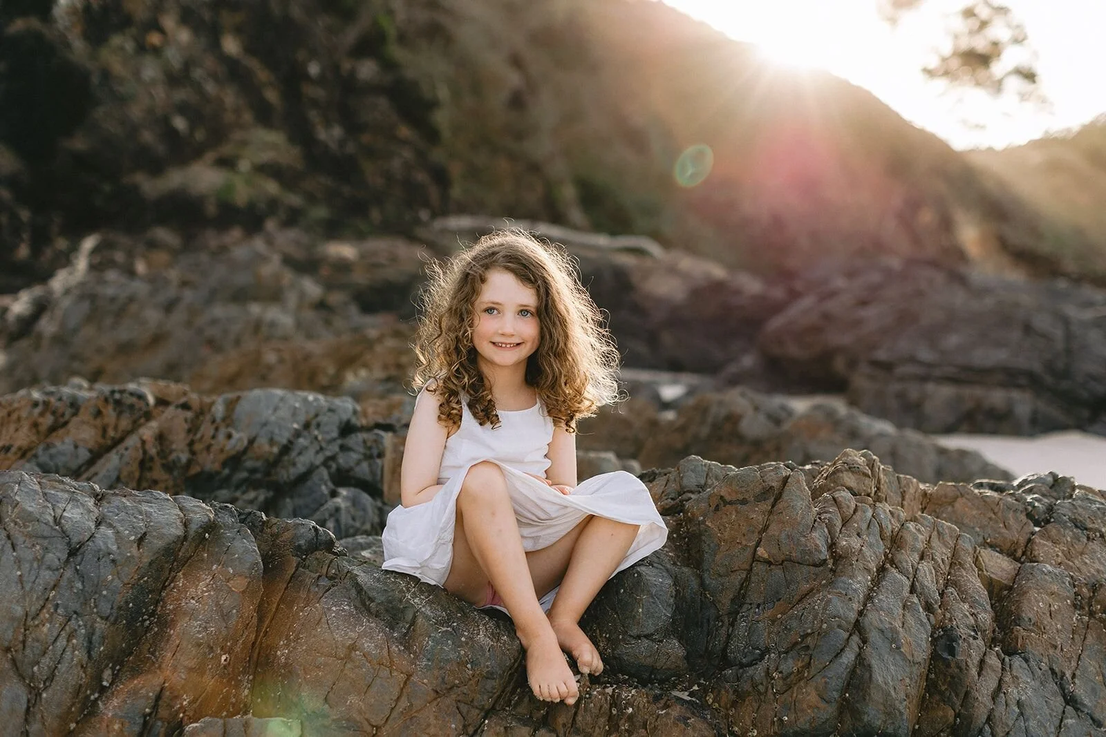 Girl in a white dress sitting on the rocks at Wategos with golden sun flaring behind her