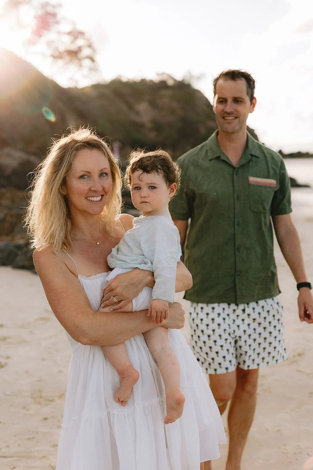 Mum holding her toddler son beside dad on the beach, headland glowing behind them