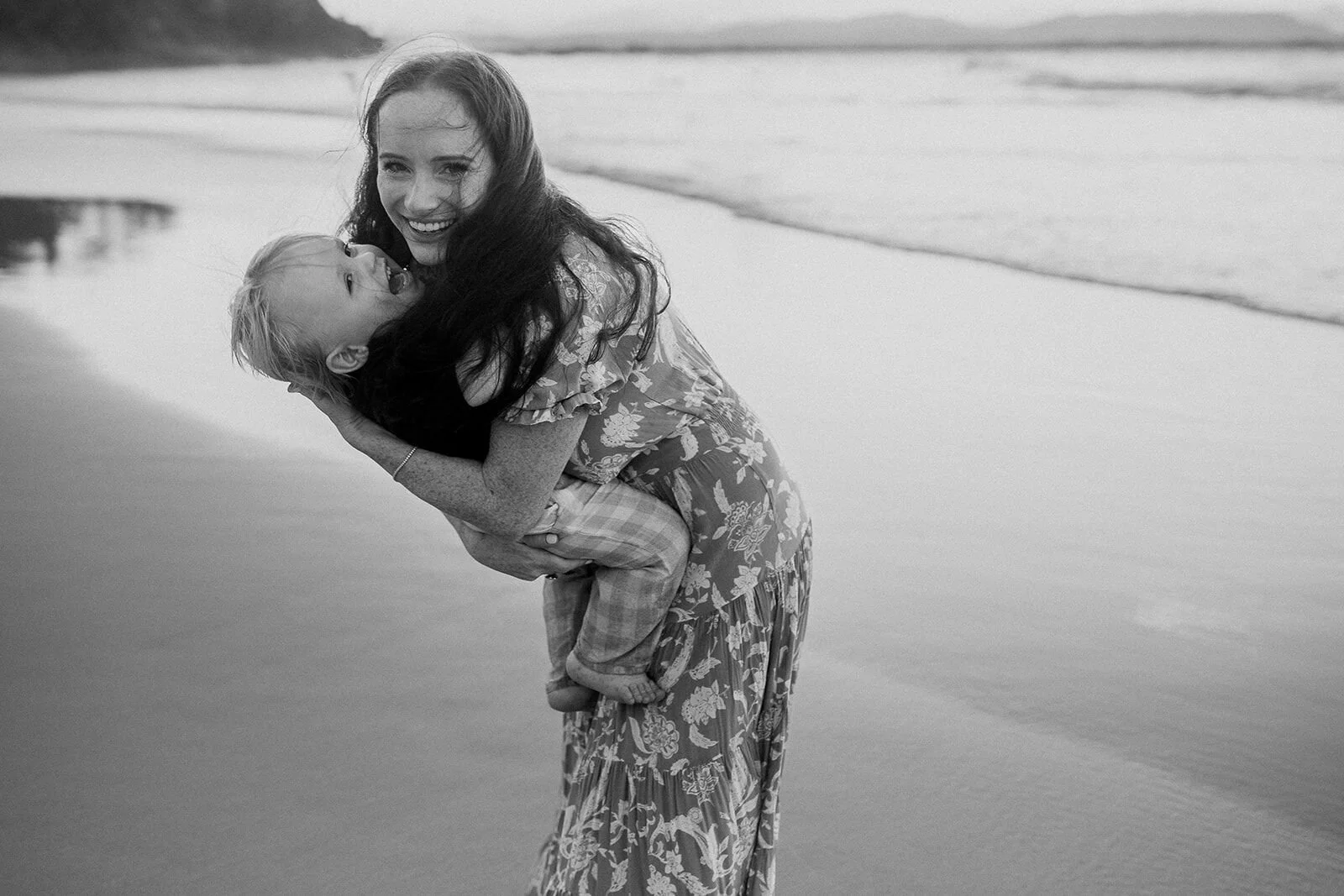 Black and white photo of mum laughing as she lifts her toddler on the beach