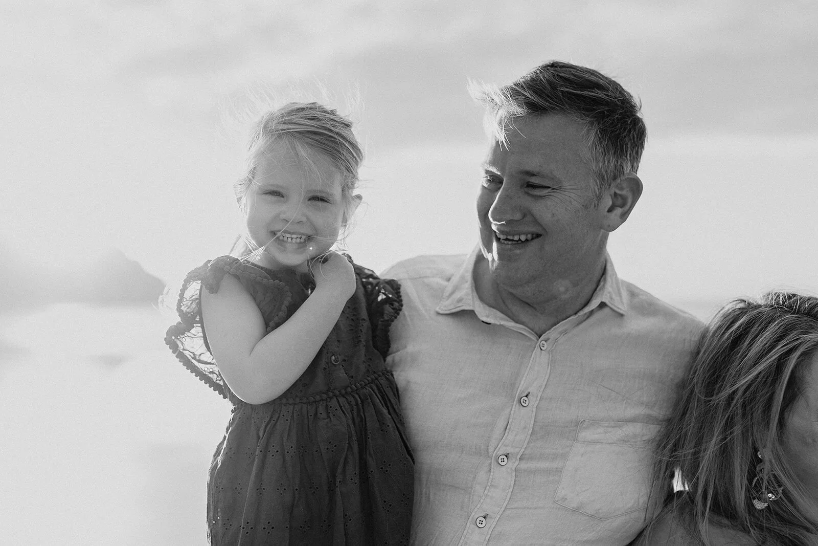Black and white photo of a little girl beaming beside her dad, who laughs on the beach