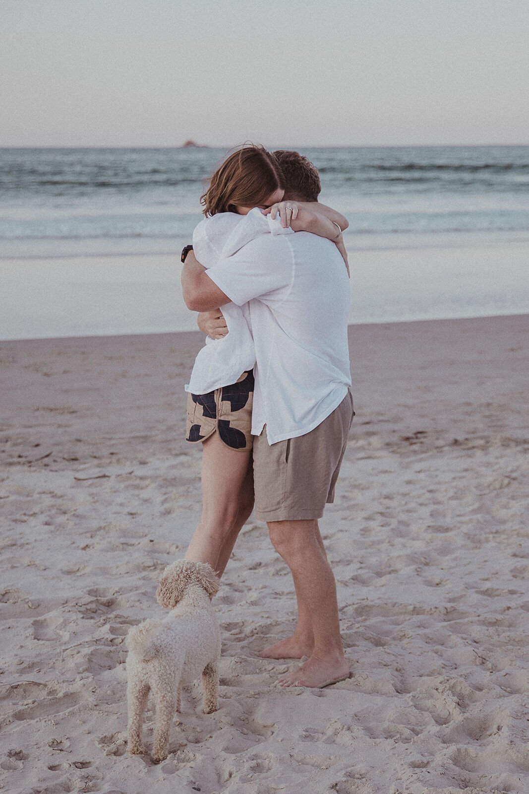 Couple hugging on the sand with their dog at their feet, Jamie and Jessica's beach marriage proposal at Belongil Beach, Byron Bay