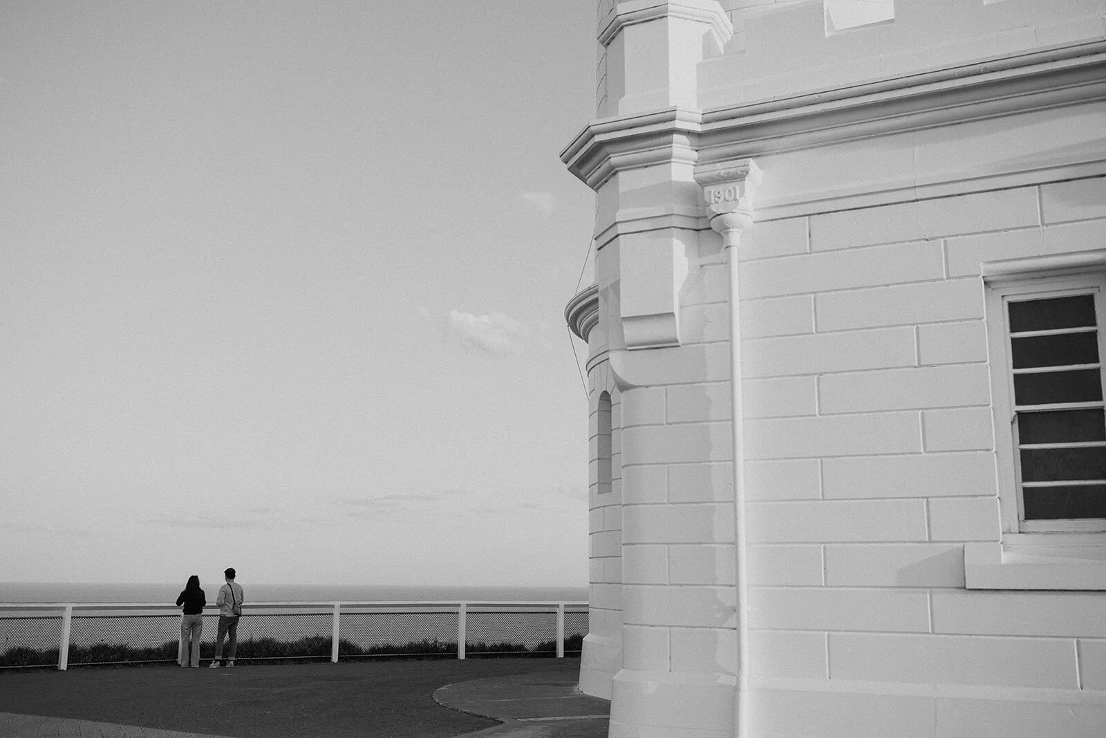 Black and white of the couple at the lookout railing beside the lighthouse, Douglas and Ying's sunset marriage proposal at Cape Byron Lighthouse, Byron Bay