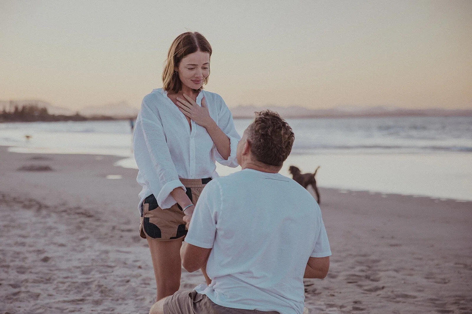 Woman with her hand on her heart as he kneels with the ring, Jamie and Jessica's beach marriage proposal at Belongil Beach, Byron Bay