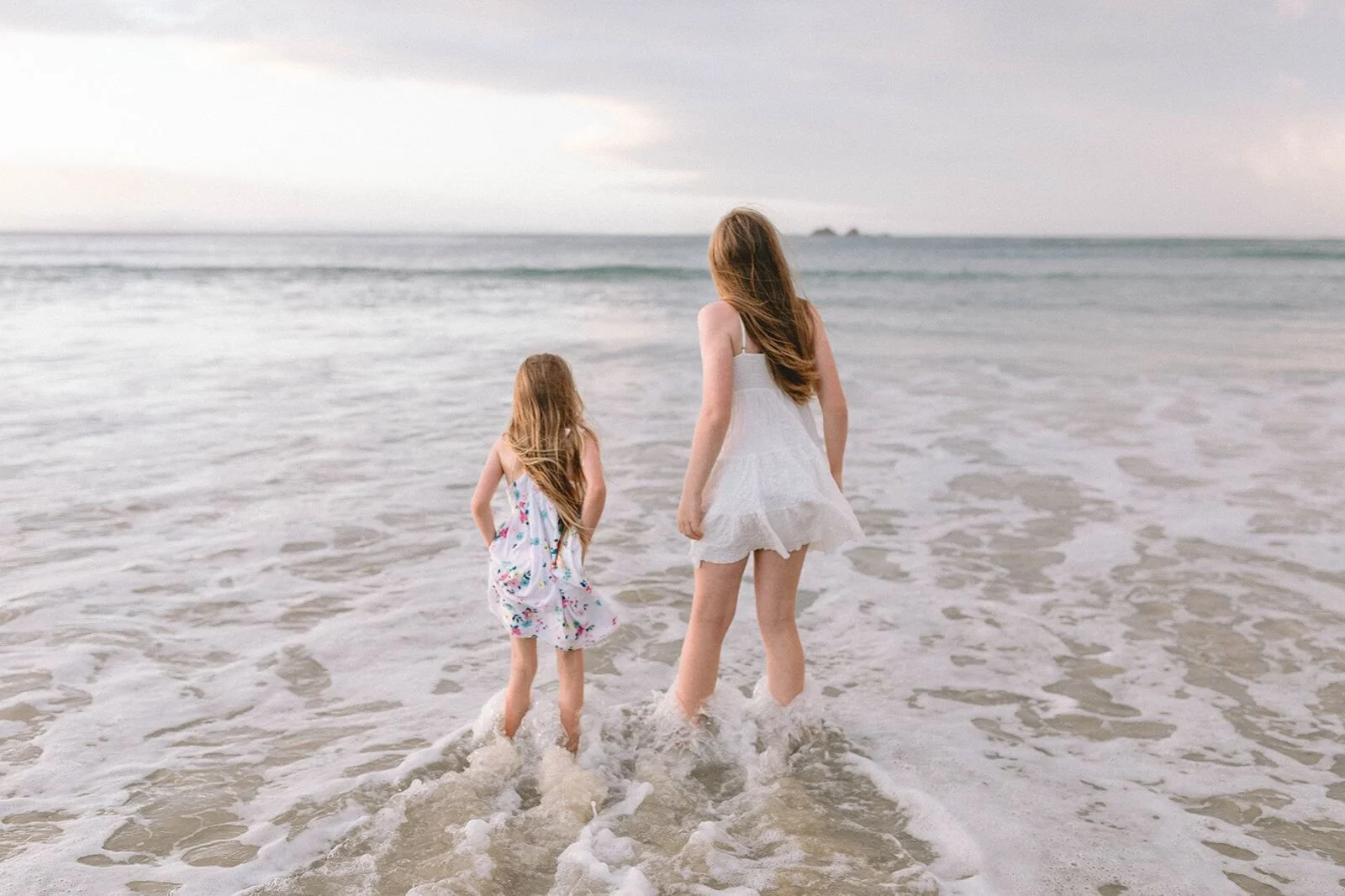 Two sisters paddling into the small waves at the water's edge, seen from behind