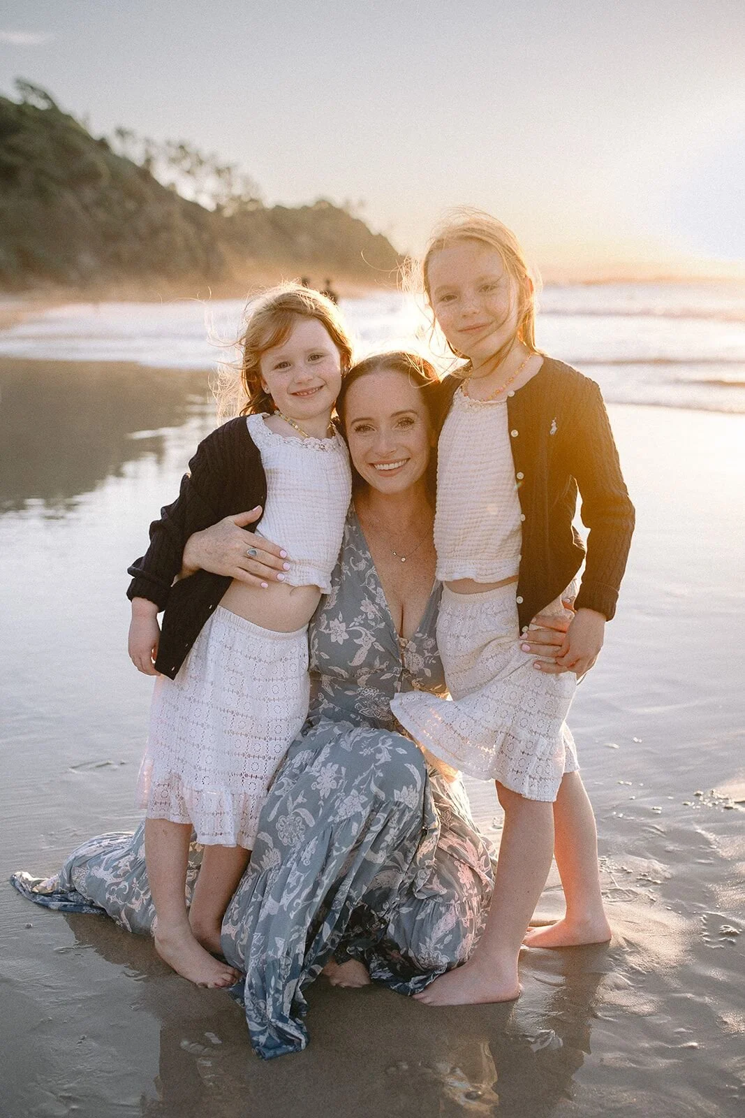 Mum cuddled in close by her two daughters on the beach with the sun glowing over the headland