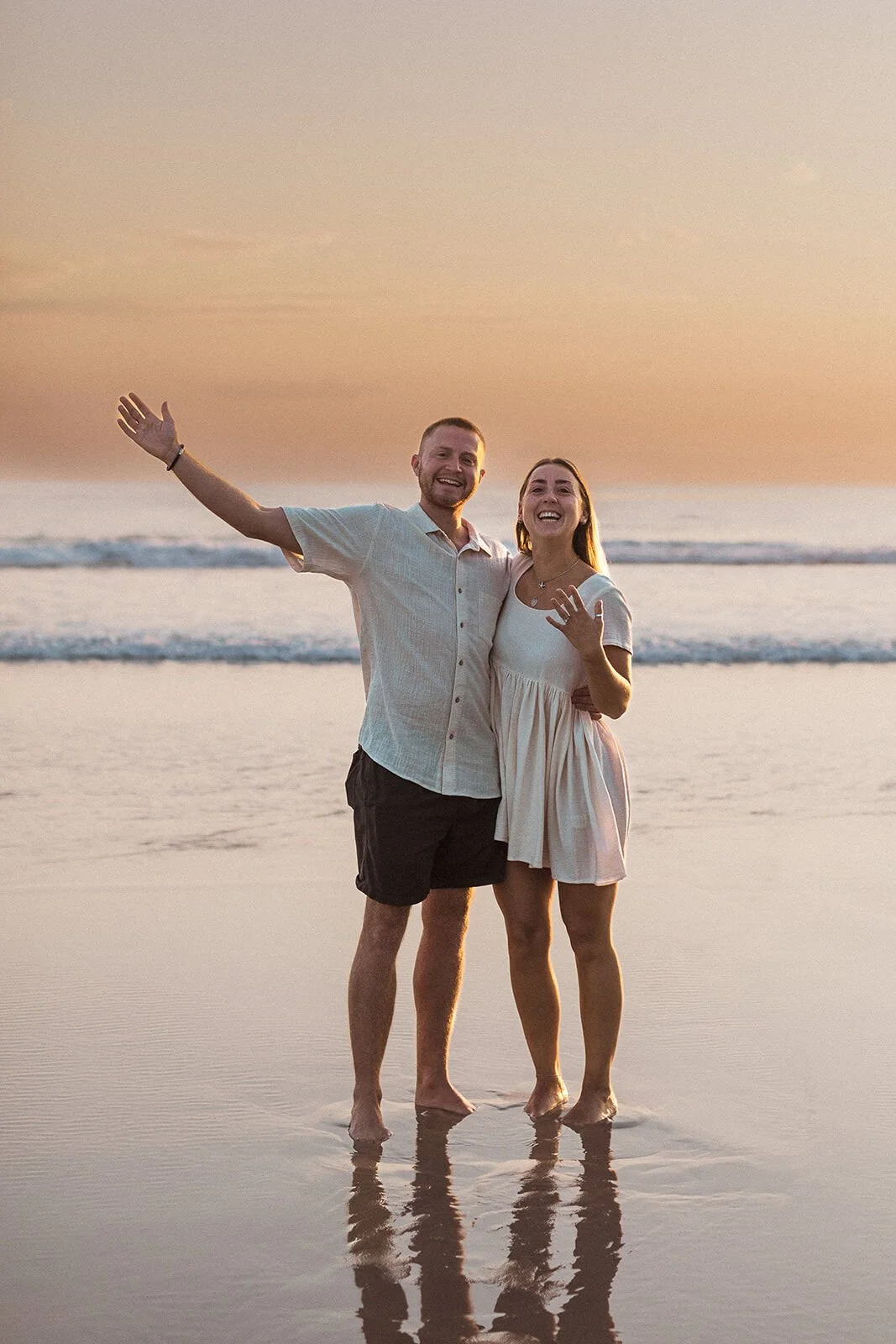 Couple celebrating, the man raising an arm as she shows her ring, Charlie and Hannah's sunrise marriage proposal at Broken Head Beach, Byron Bay
