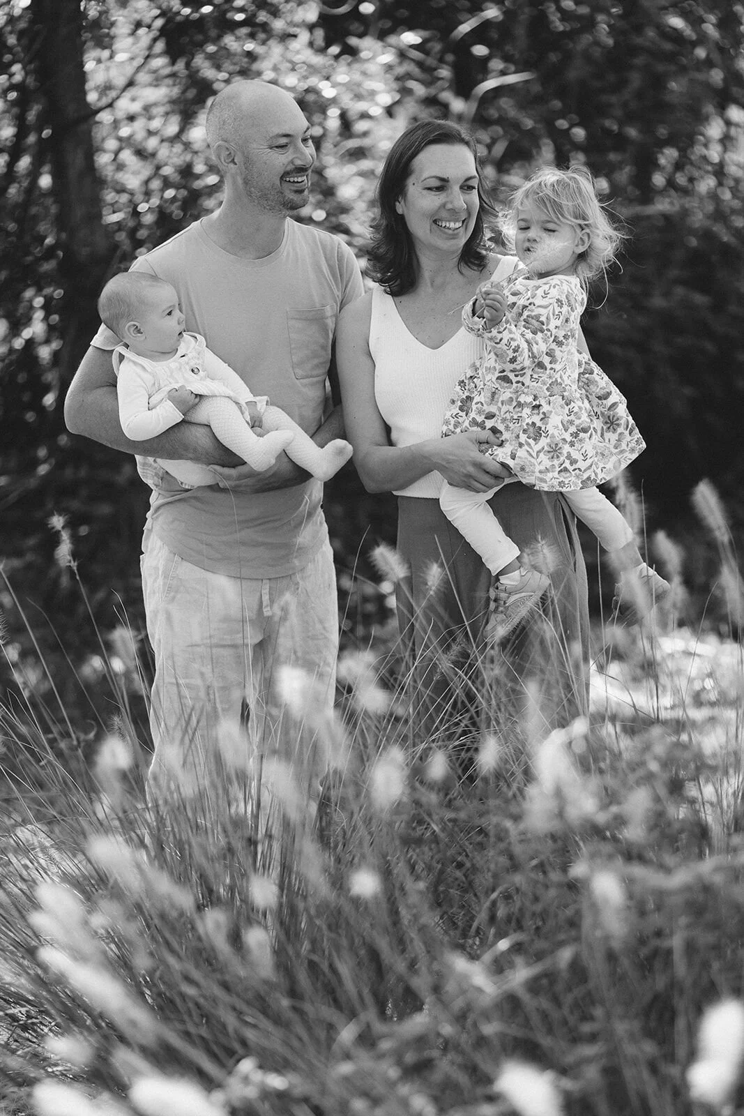 Black and white family portrait in tall grass, mum and dad each holding a daughter