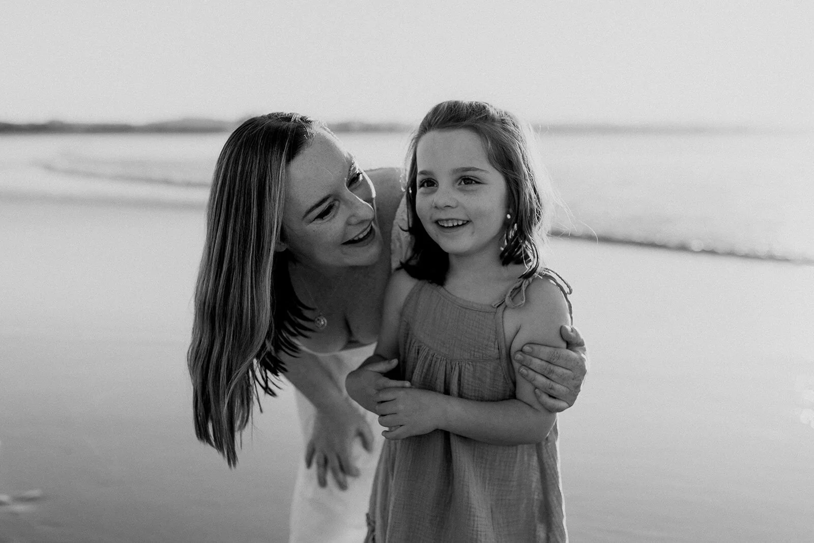 Black and white photo of mum leaning down to hug her smiling daughter on the beach